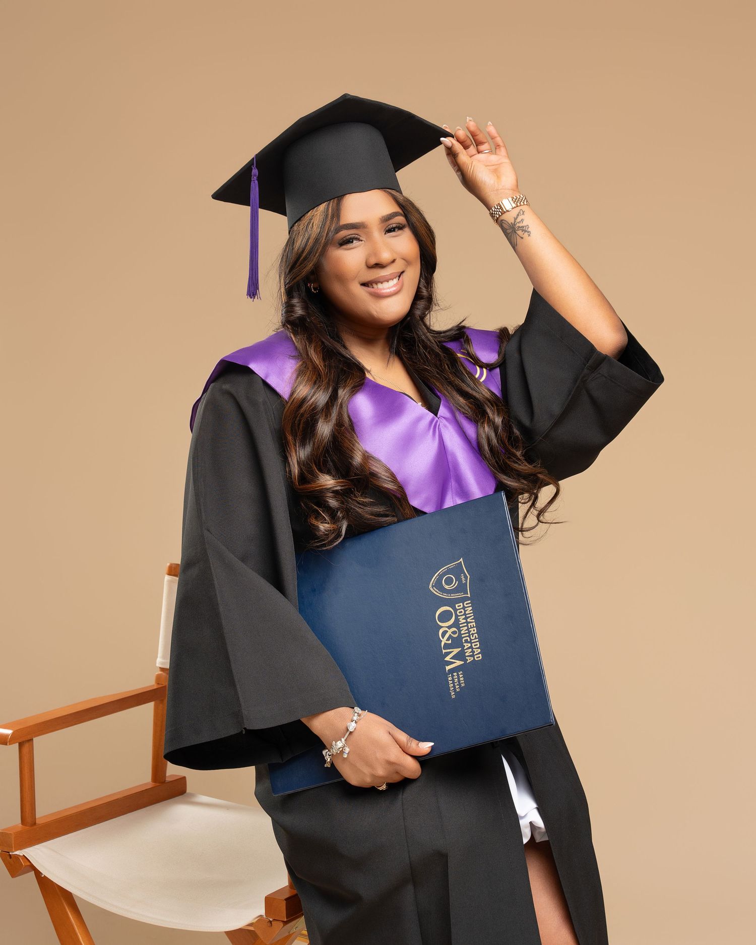 Smiling graduate in black cap and gown holding diploma, seated in wooden chair against neutral background.