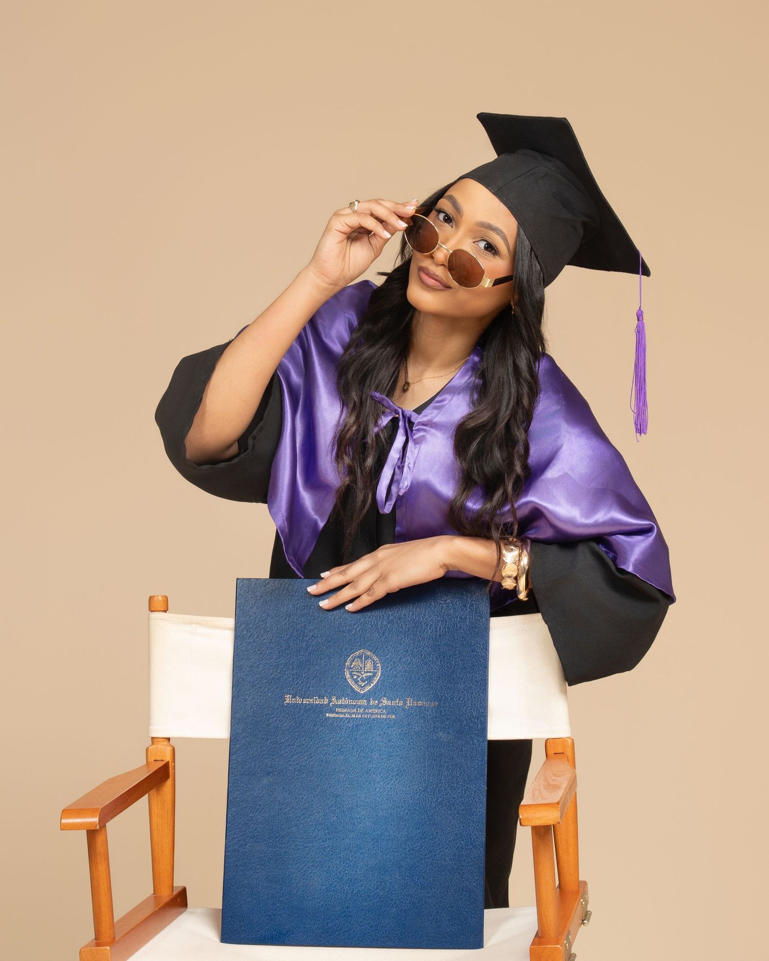Smiling graduate in black cap and gown holding diploma, seated in wooden chair against neutral background.