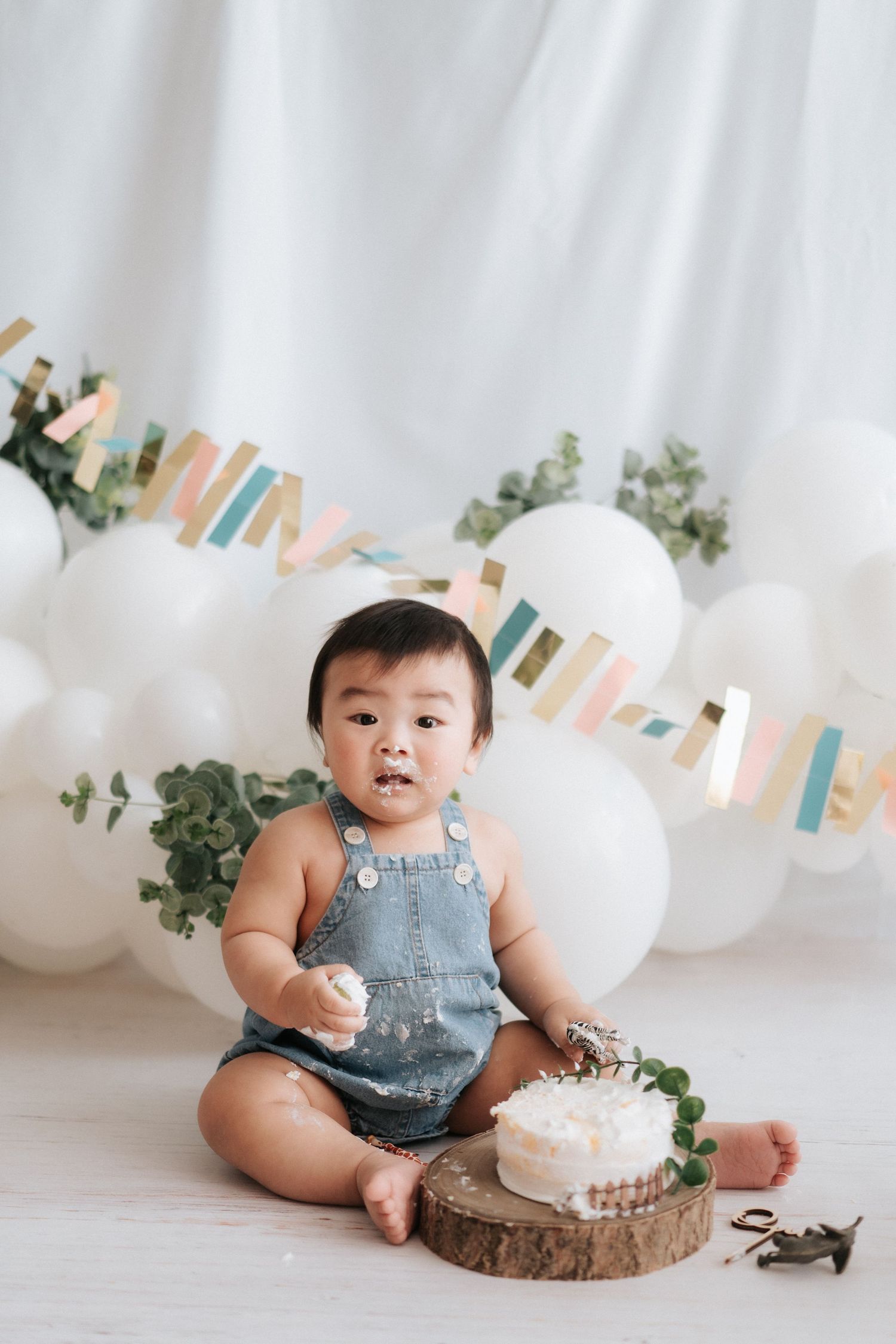 A baby in denim overalls sits on a wooden slice with birthday decorations, white balloons and pastel banners in background.