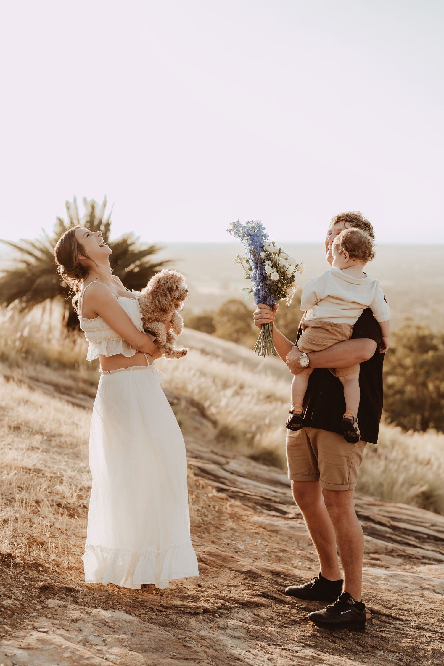A family in casual and formal attire stands together on a hilltop during golden hour holding wildflowers.