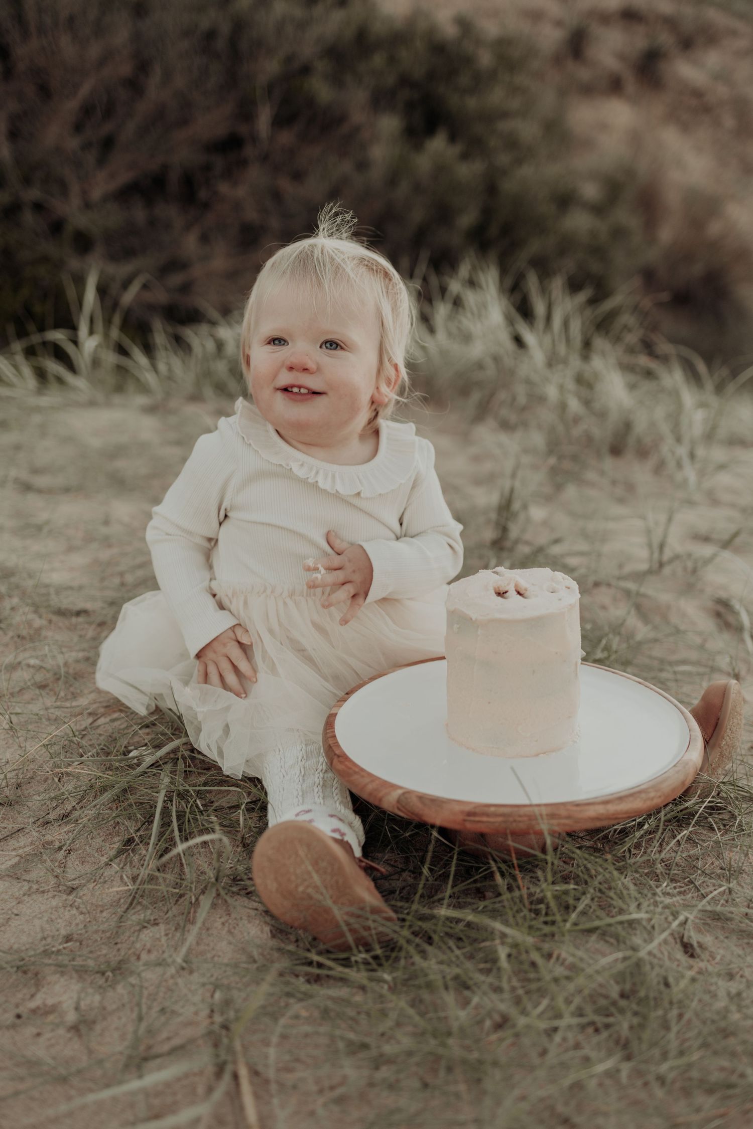 A smiling baby girl in a white dress sits on sandy ground next to a small cake on a wooden board.