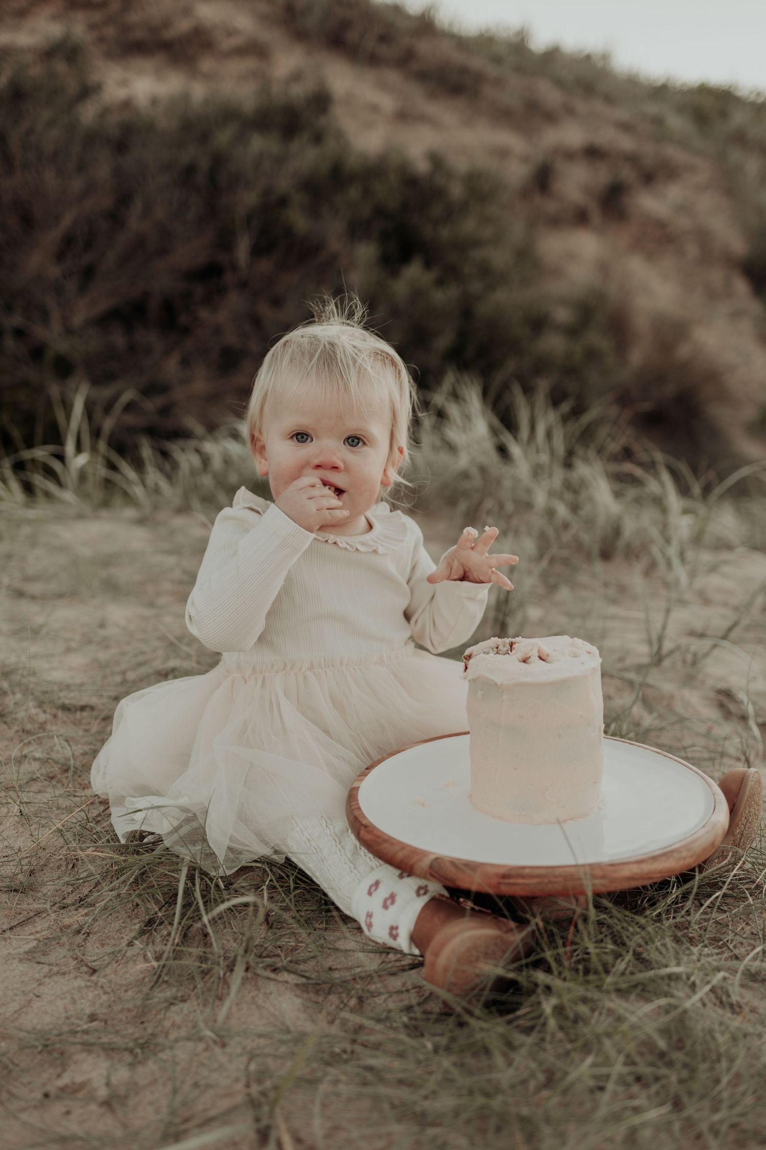 Baby girl in white dress eating her first birthday smash cake outdoors on sandy dunes.