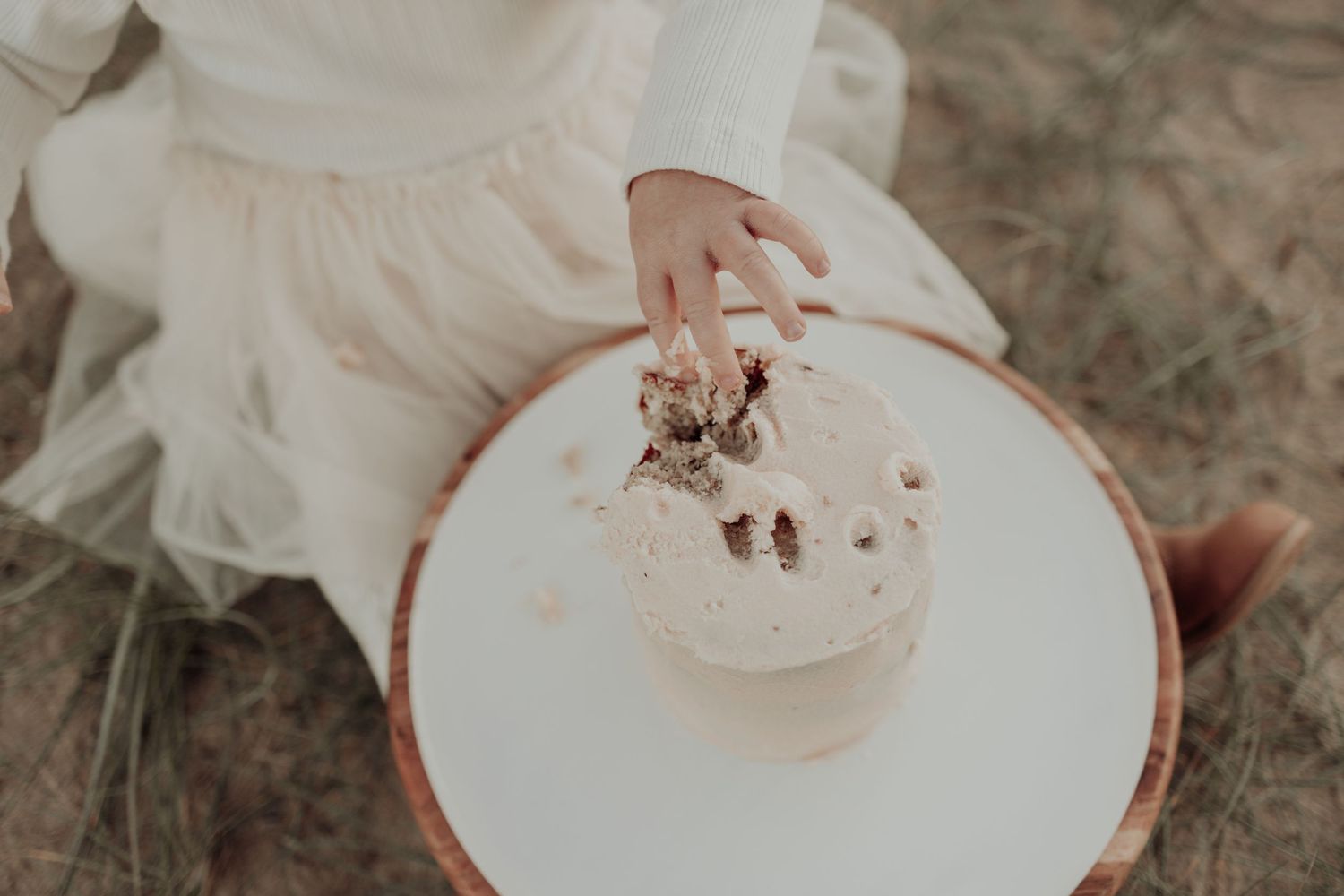 A child in a white tulle dress reaches down to touch a cream cake on a small round table outdoors.