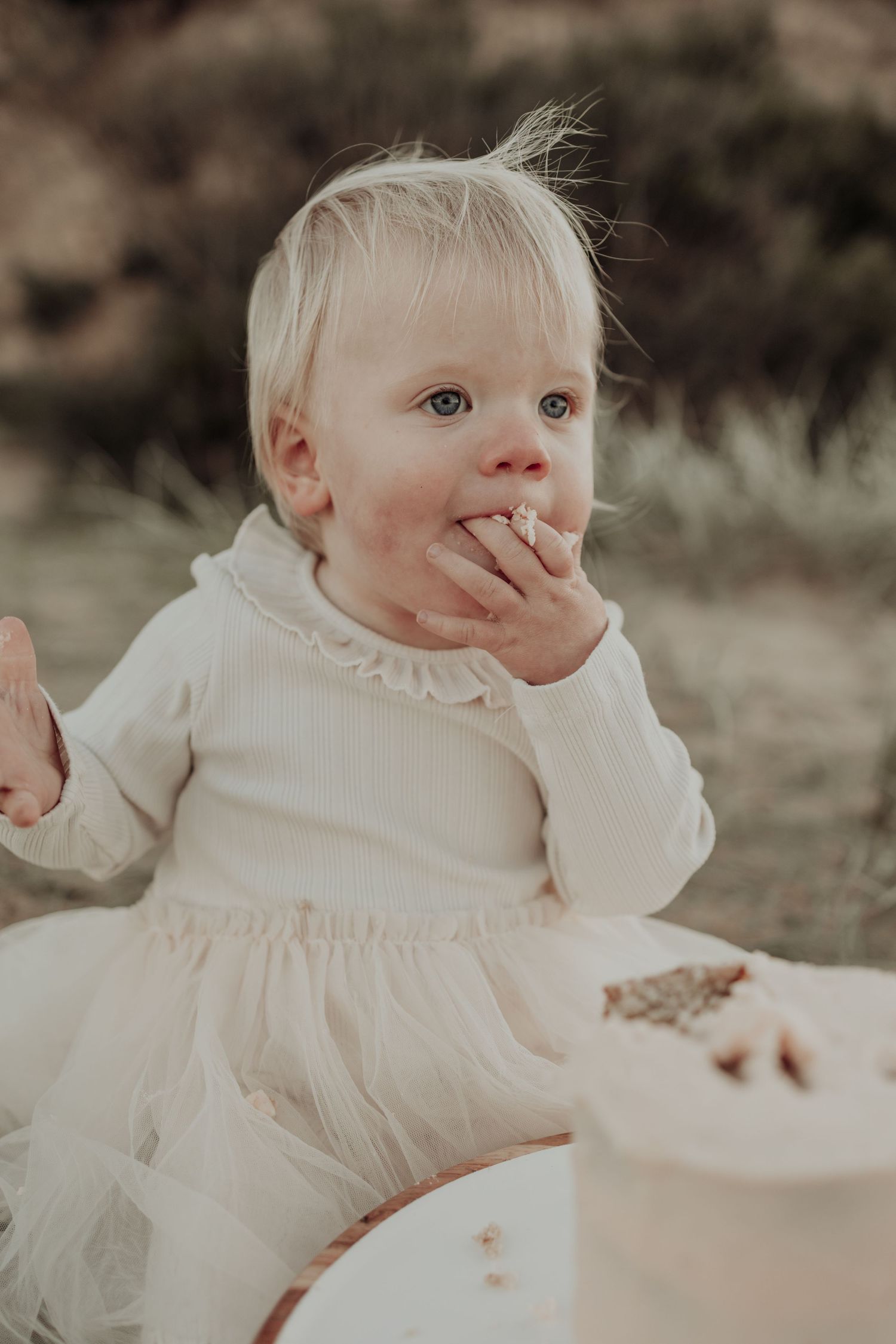 Adorable blonde baby girl in white dress eating cake outdoors during first birthday smash cake photo session.