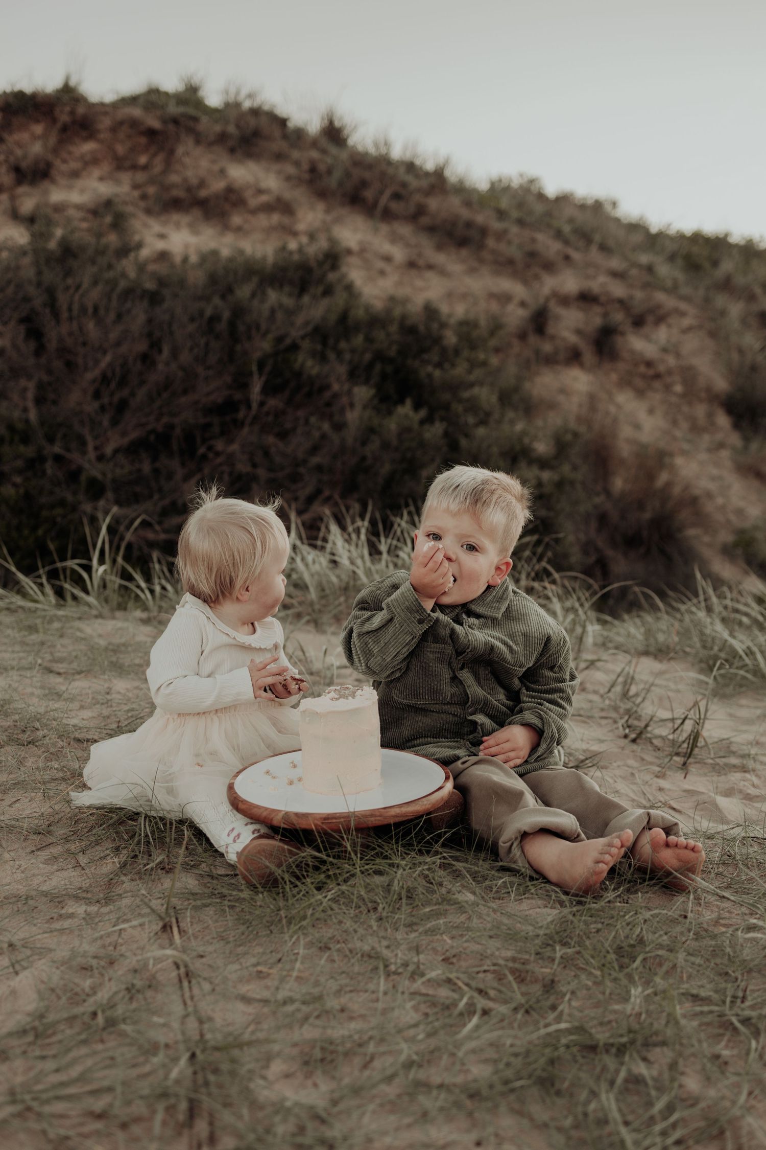 Two toddlers sit on sandy dunes sharing a small cake outdoors, dressed in white and green outfits.