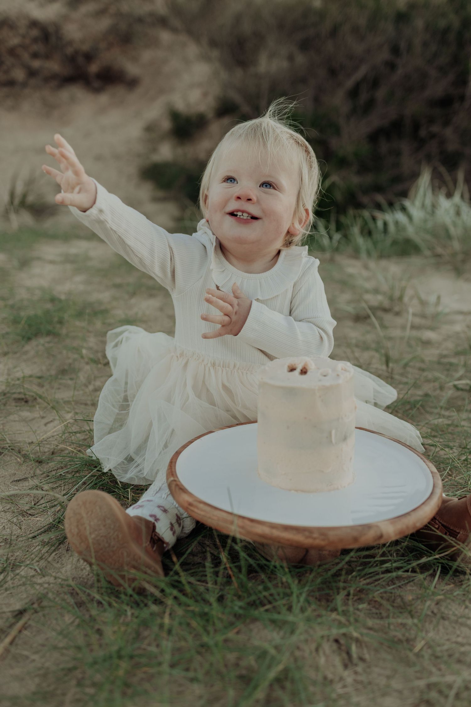 Joyful toddler girl in white dress sitting outdoors with a small white birthday cake on a wooden board.