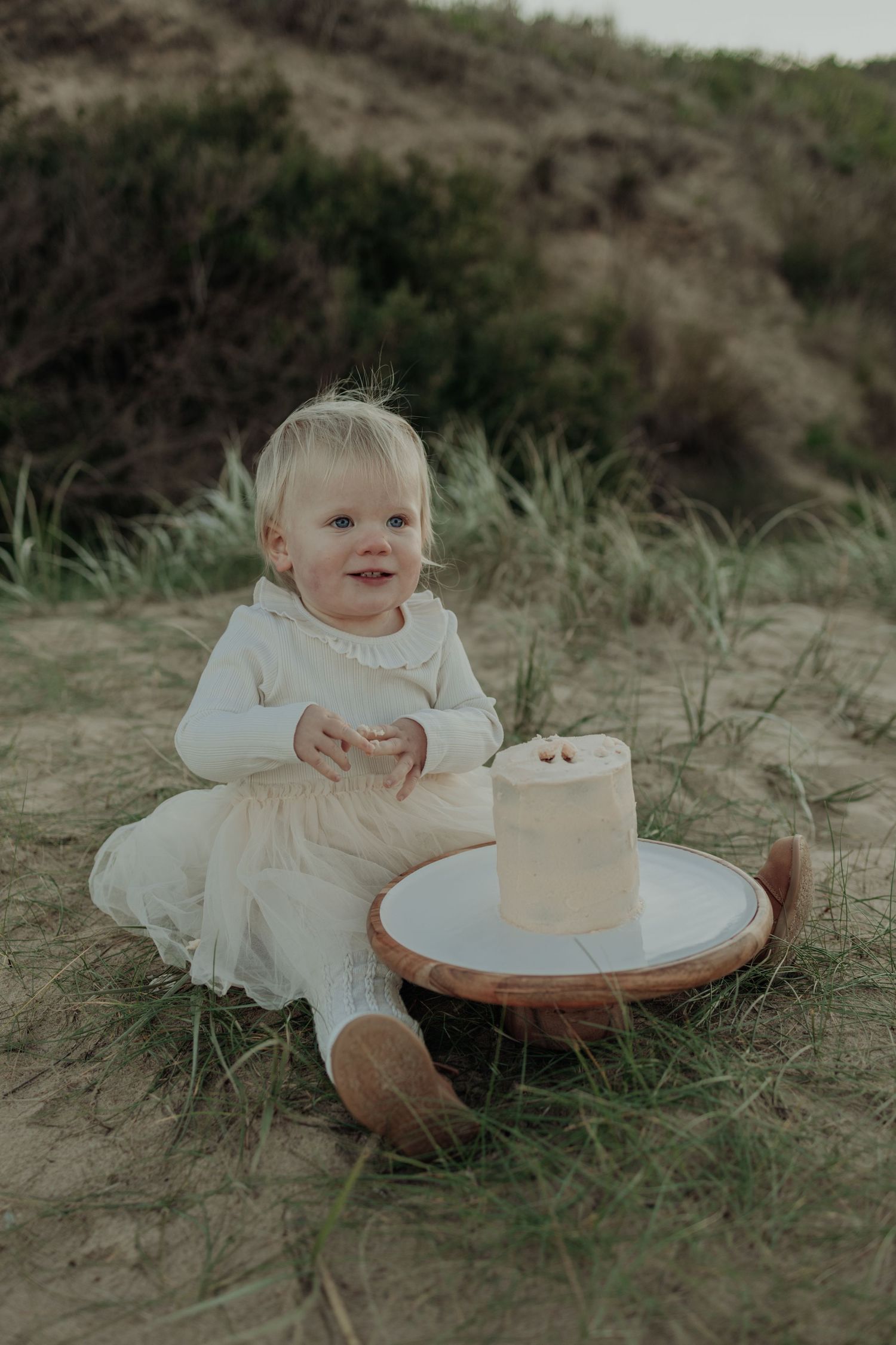 Smiling baby girl in white dress sitting on sandy beach dunes with a small cake on a wooden tray.