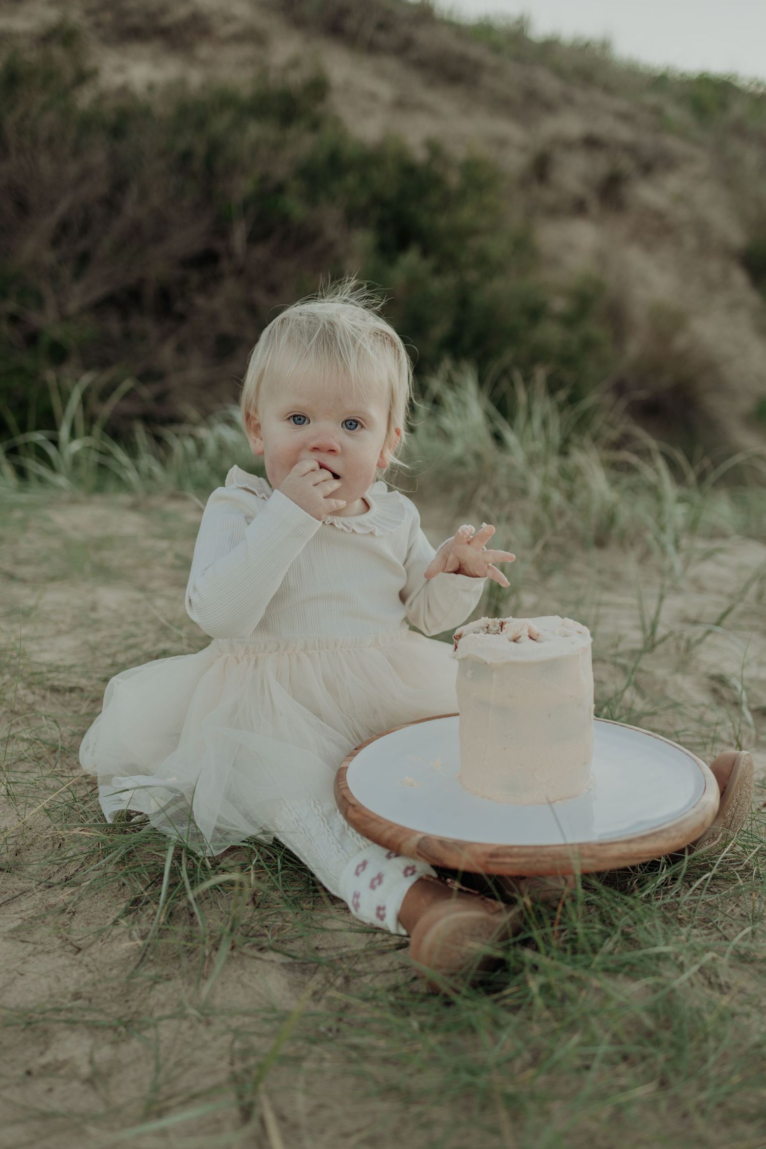 Baby girl in white dress eating smash cake outdoors on sandy beach dunes for first birthday celebration.