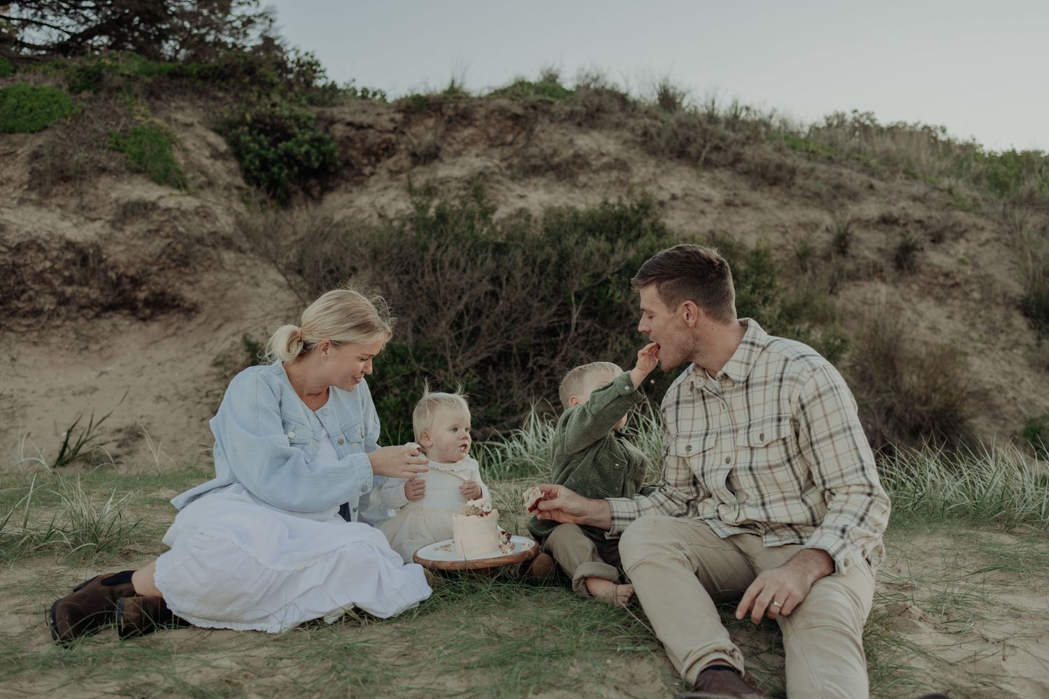 Family of three enjoying a beach picnic in sandy dunes, parents smiling at baby during outdoor celebration.