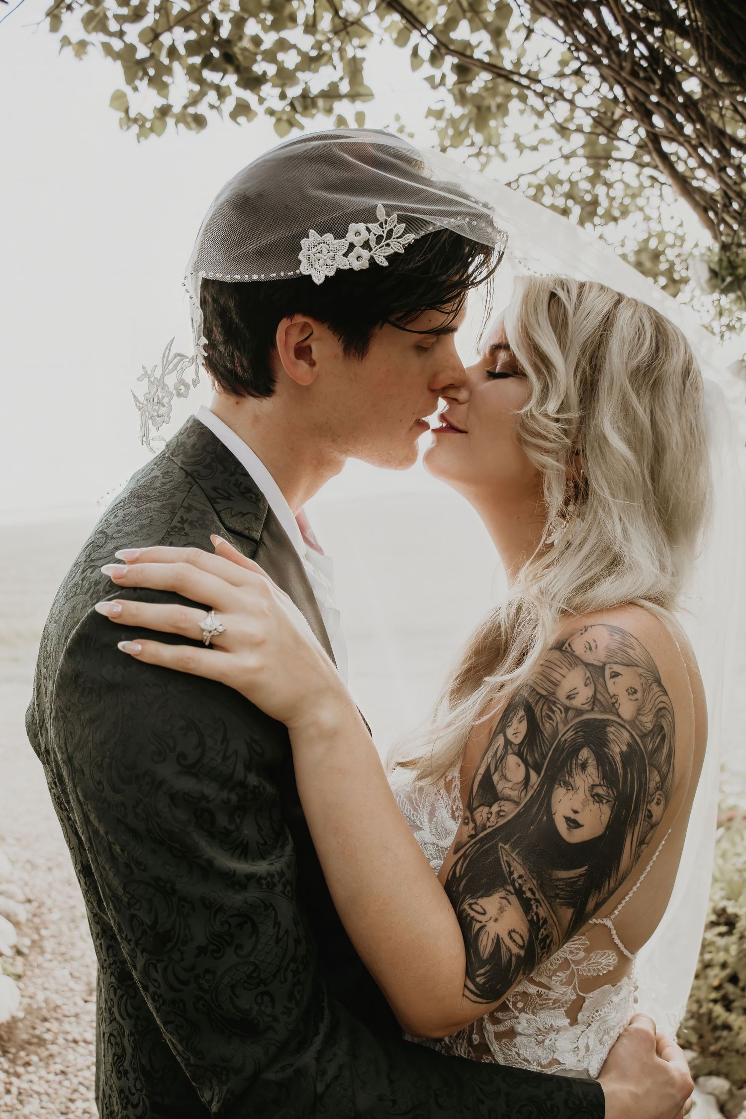 Bride and groom sharing romantic kiss under tree, bride with floral sleeve tattoo in lace gown.