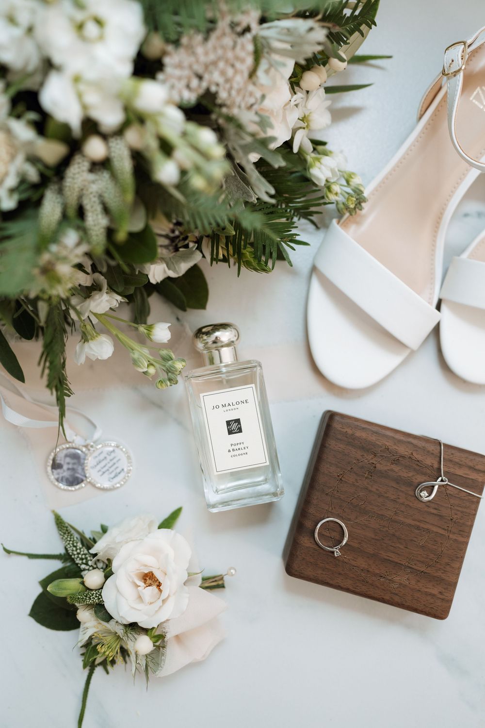 Wedding details flatlay with white shoes, perfume bottle, wooden ring box and floral arrangements.