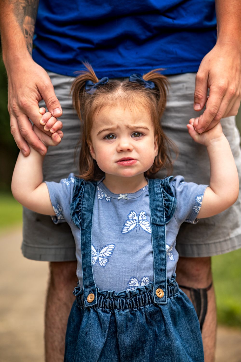 Child in denim overalls and blue shirt with butterfly pattern holds hands with adult in casual summer setting.