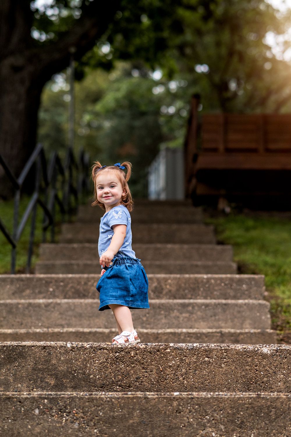 A toddler in denim overalls sits playfully on outdoor concrete steps during golden hour.