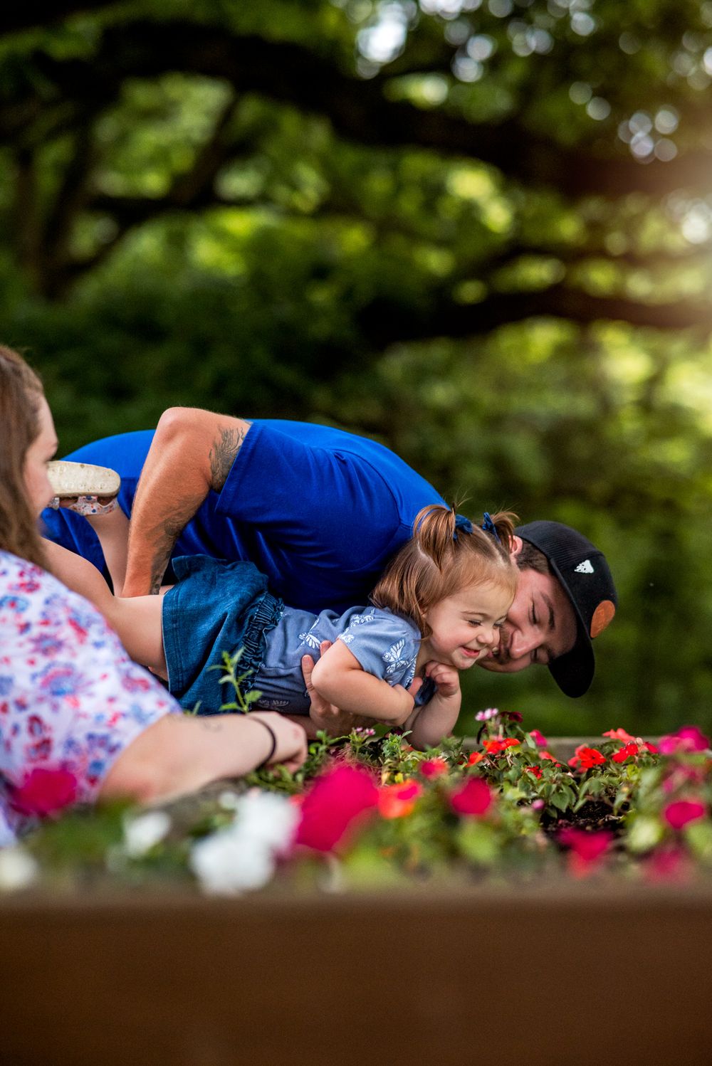 A couple in blue outfits share a tender moment lying together among red flowers in a garden setting.