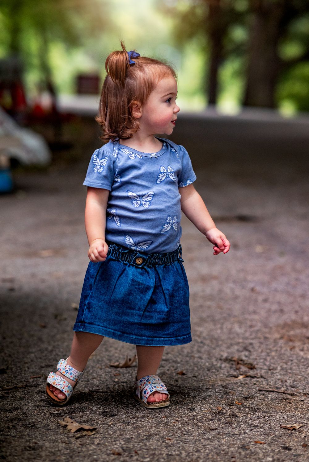 A toddler wearing a blue top and denim skirt explores a dirt path in a wooded outdoor setting.