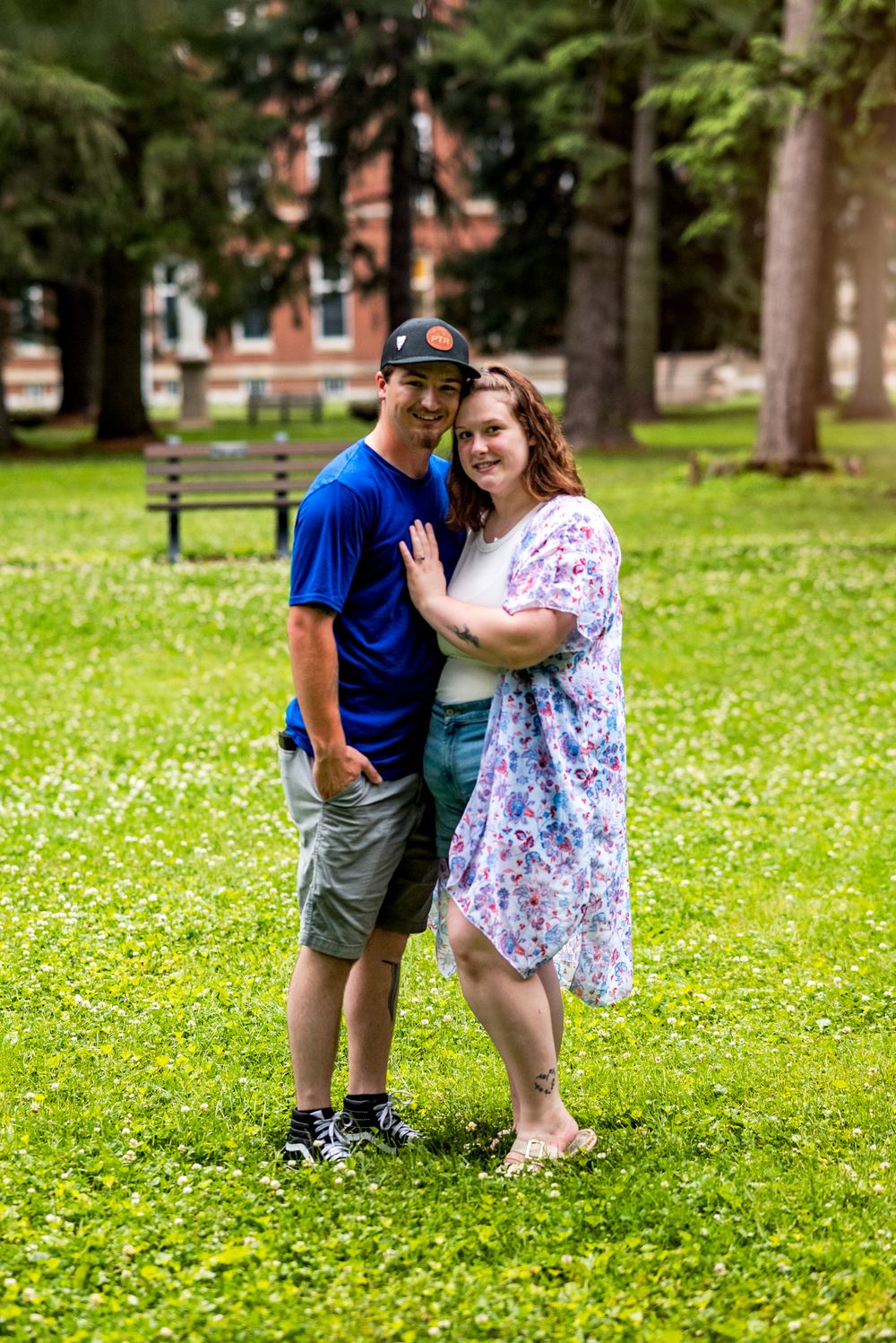 A couple poses together on a grassy lawn with trees and a brick building in the background.