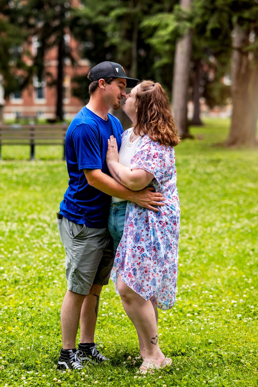 A couple in a park share a tender moment while standing on grass dotted with small white flowers.