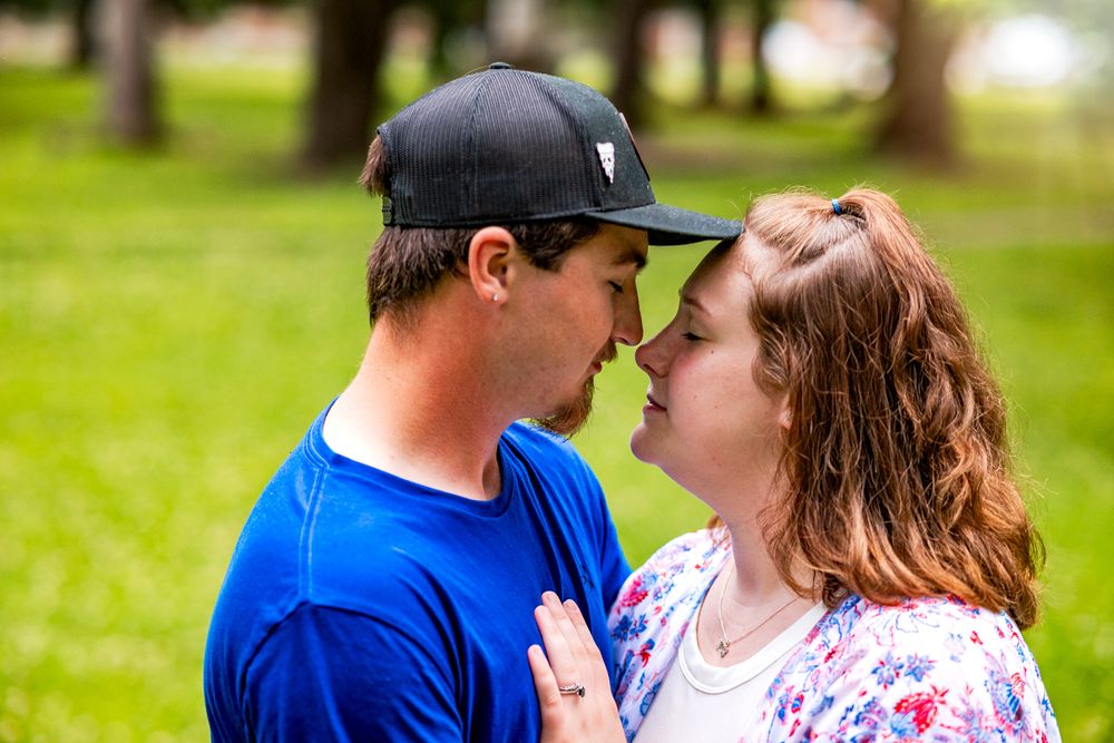A couple in blue and pink clothing share a tender moment outdoors in a park setting.