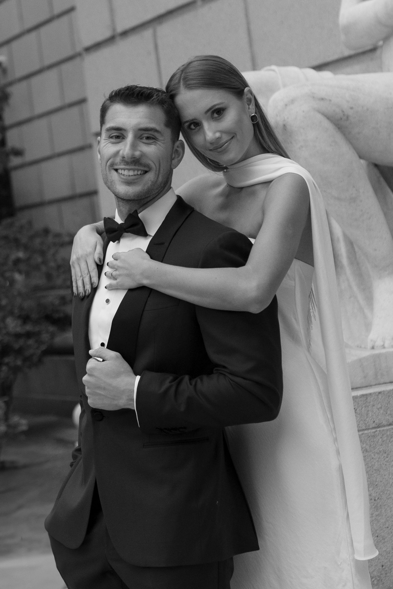 Black and white wedding photo of a bride in white dress embracing groom in tuxedo from behind during formal portraits.