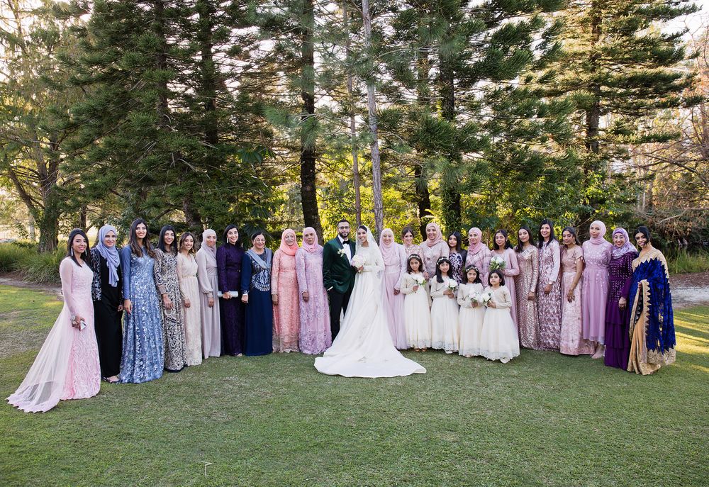 Large wedding party group photo on green lawn surrounded by tall trees in outdoor garden setting.