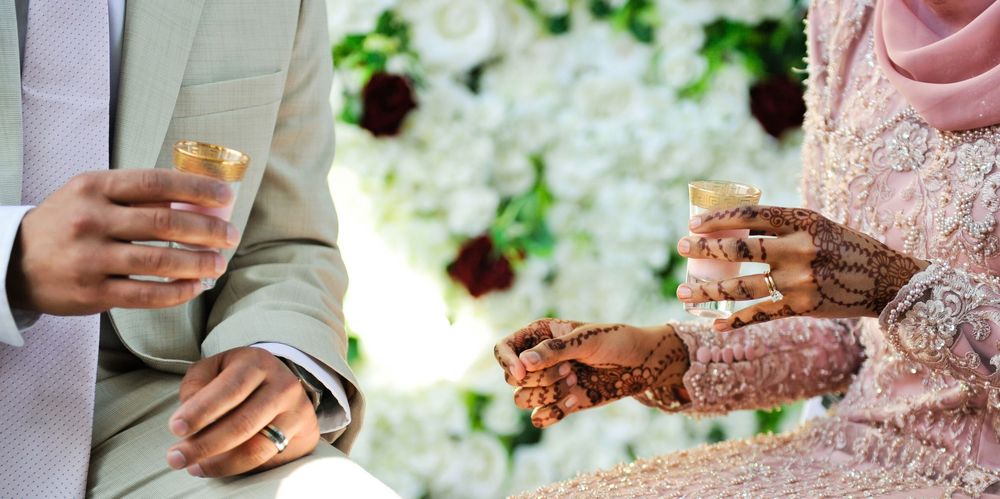 Muslim wedding couple exchanging rings with henna decorated hands and white floral backdrop.