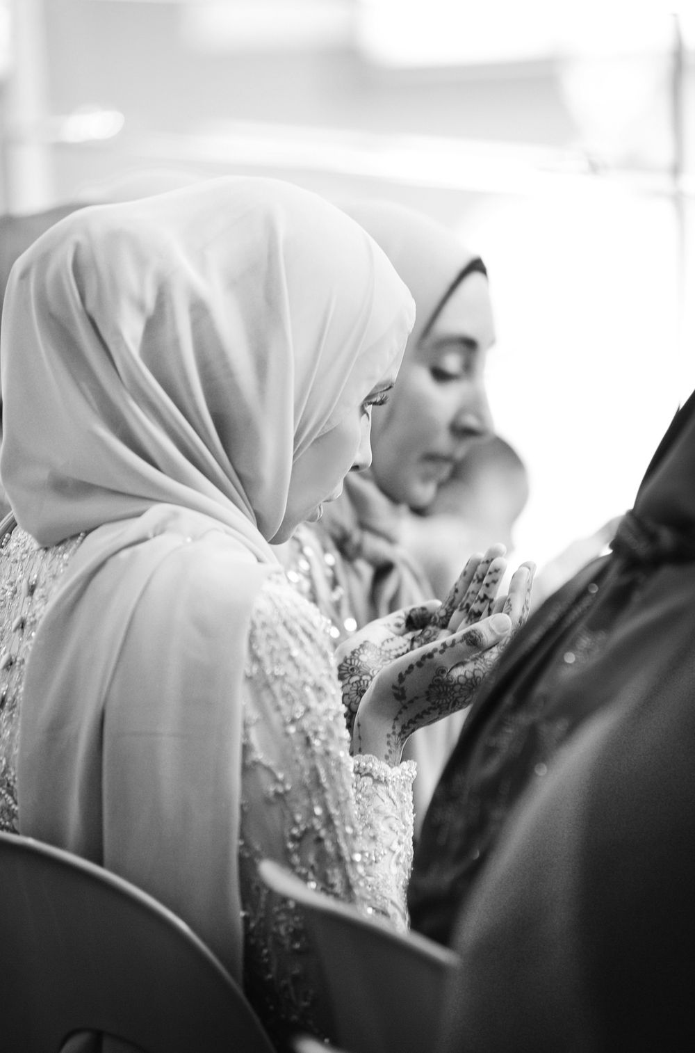 Black and white photo of Muslim bride in hijab praying with henna decorated hands at wedding.