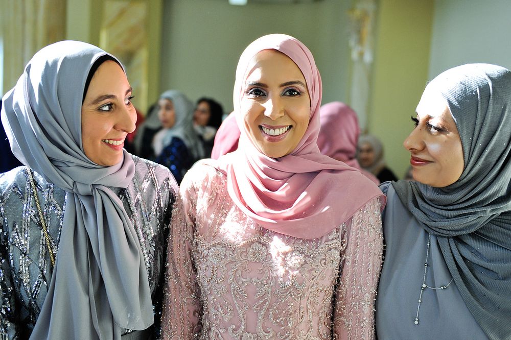 Three Muslim women in elegant hijabs and lace dresses smiling together at a wedding celebration.
