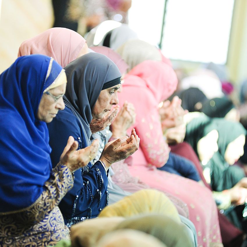Muslim women in colorful hijabs seated and praying together at a wedding ceremony.