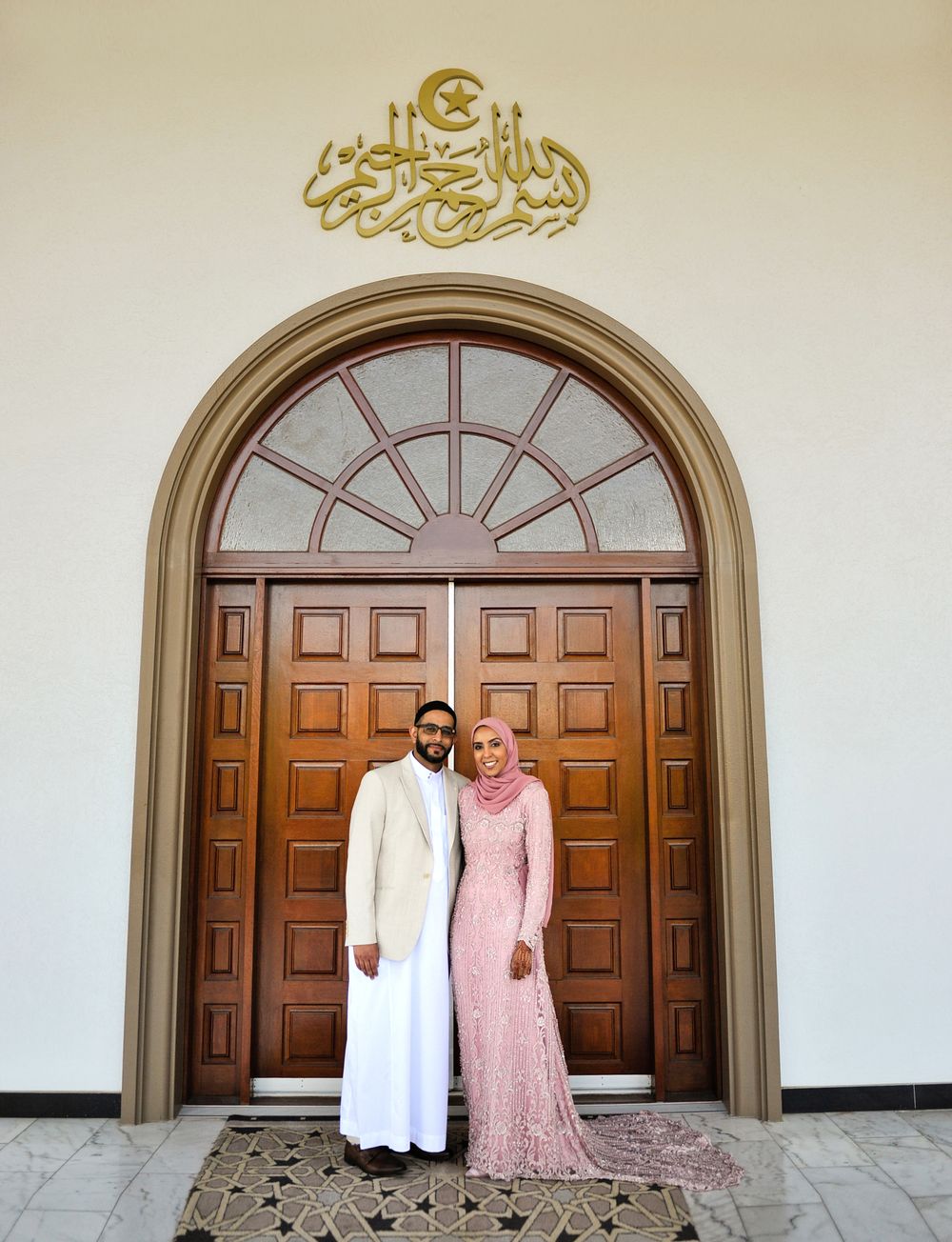 Muslim bride and groom posing before ornate mosque entrance with Arabic calligraphy above doorway.