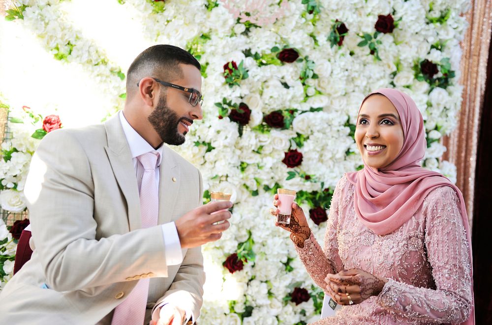 Couple in elegant wedding attire toasting with drinks in front of a white and red floral wall backdrop.