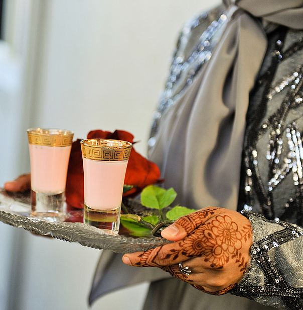 Ornate gold-rimmed glasses with pink drinks on a silver tray held by hands adorned with henna at a wedding.