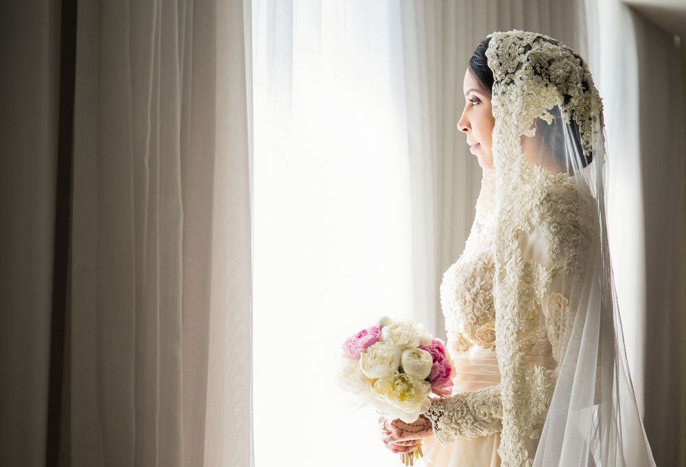 Bride in lace wedding dress and floral hijab holding bouquet while gazing out a sunlit window.