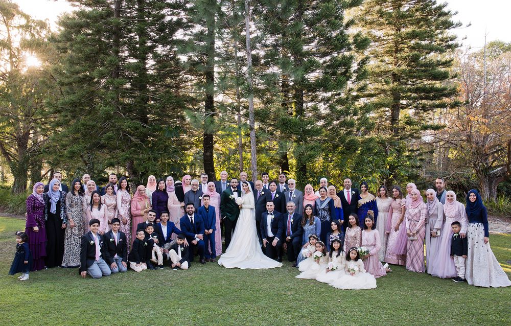 Large Muslim wedding party group portrait at sunset in a lush green outdoor garden with tall trees.