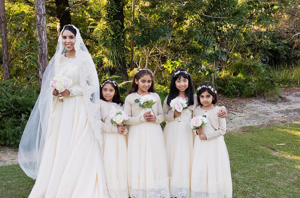 Bride in white lace gown and veil posing with four flower girls in matching cream dresses outdoors.