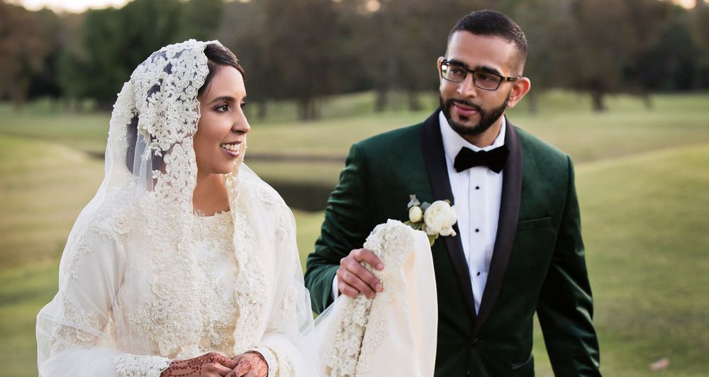 Muslim bride in white lace gown and veil walking with groom in green velvet tuxedo on golf course.