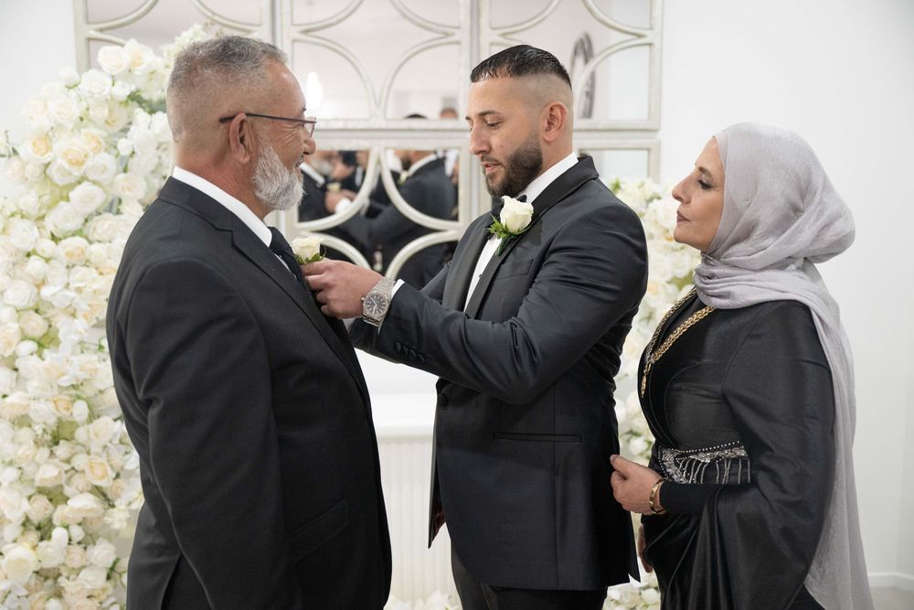 Groom in black tuxedo receiving boutonniere from parents in front of white floral wedding arch.