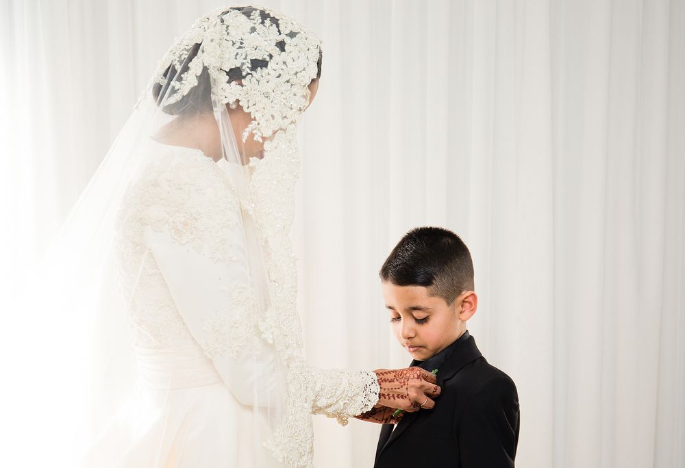 Bride in white lace wedding dress and veil pinning boutonniere on young boy in black suit.