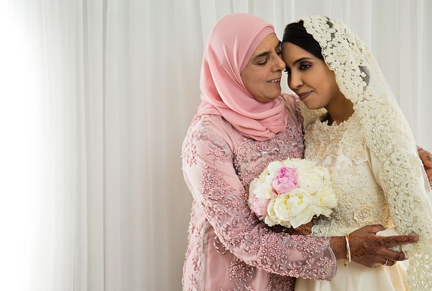 Muslim bride in white lace wedding dress embracing woman in pink hijab holding a floral bouquet.