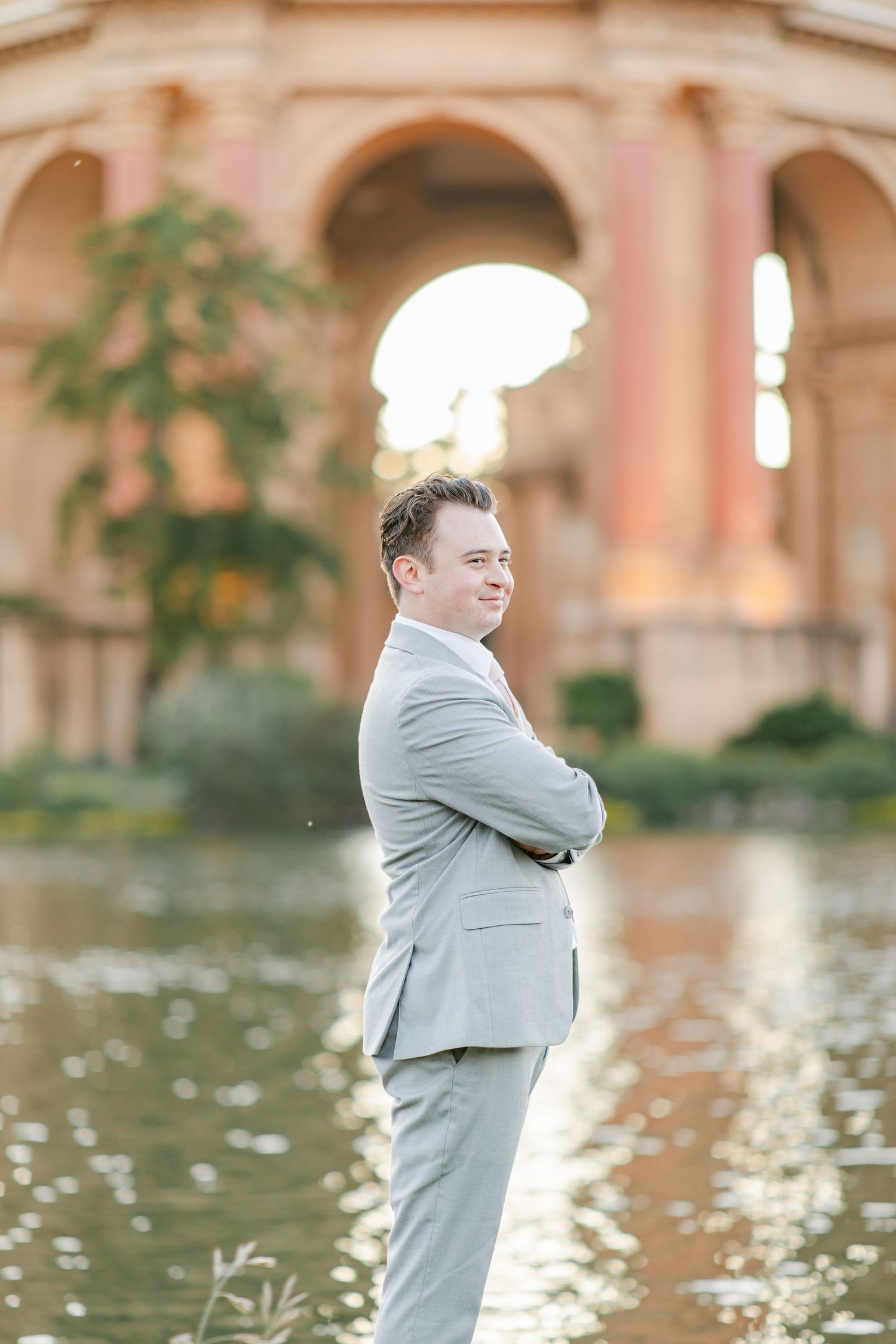 A person in a light gray suit stands near a reflective pond with ornate architecture in the background.