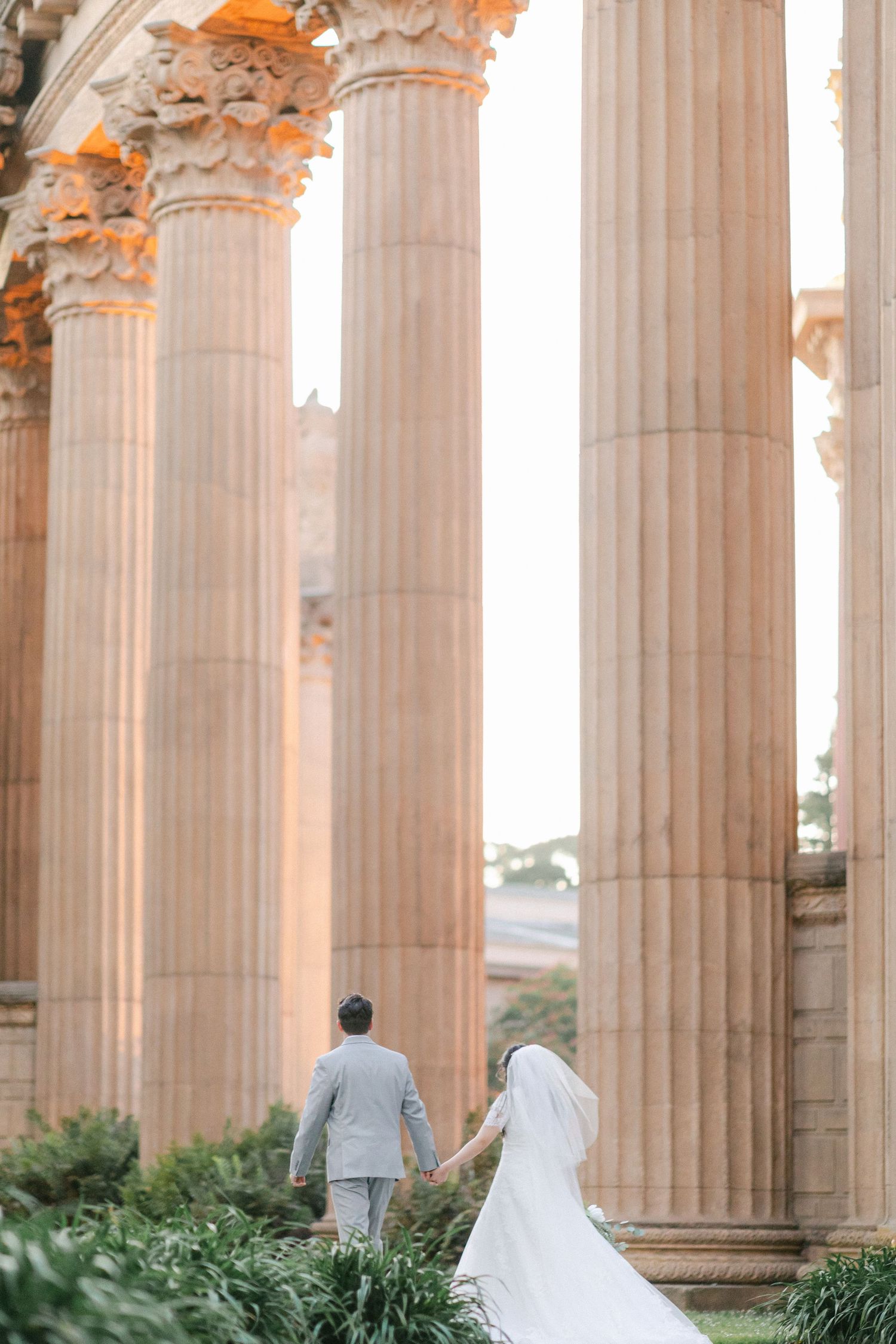 A couple in wedding attire walk among grand classical columns at sunset, creating an elegant architectural backdrop.