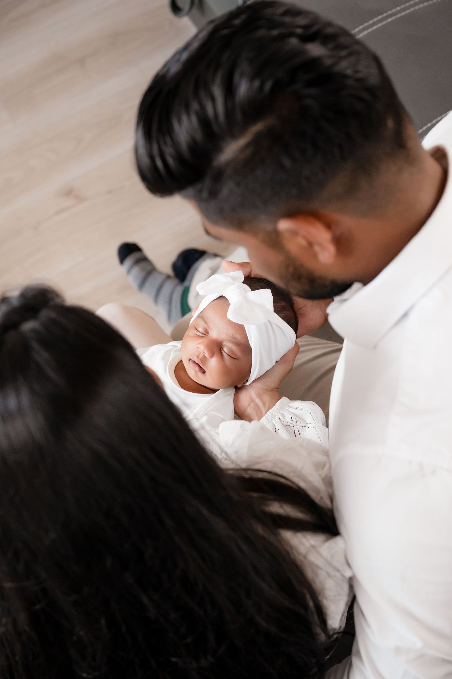 Parents lovingly gaze at their newborn baby wearing a white bow headband while resting on white bedding.