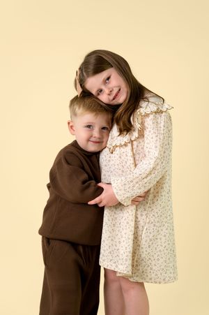 Two children in brown and cream outfits hugging and smiling against a beige background during a studio photoshoot.