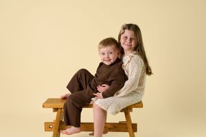 Two figures in brown and cream colored clothing sit together on a wooden bench in a studio setting.