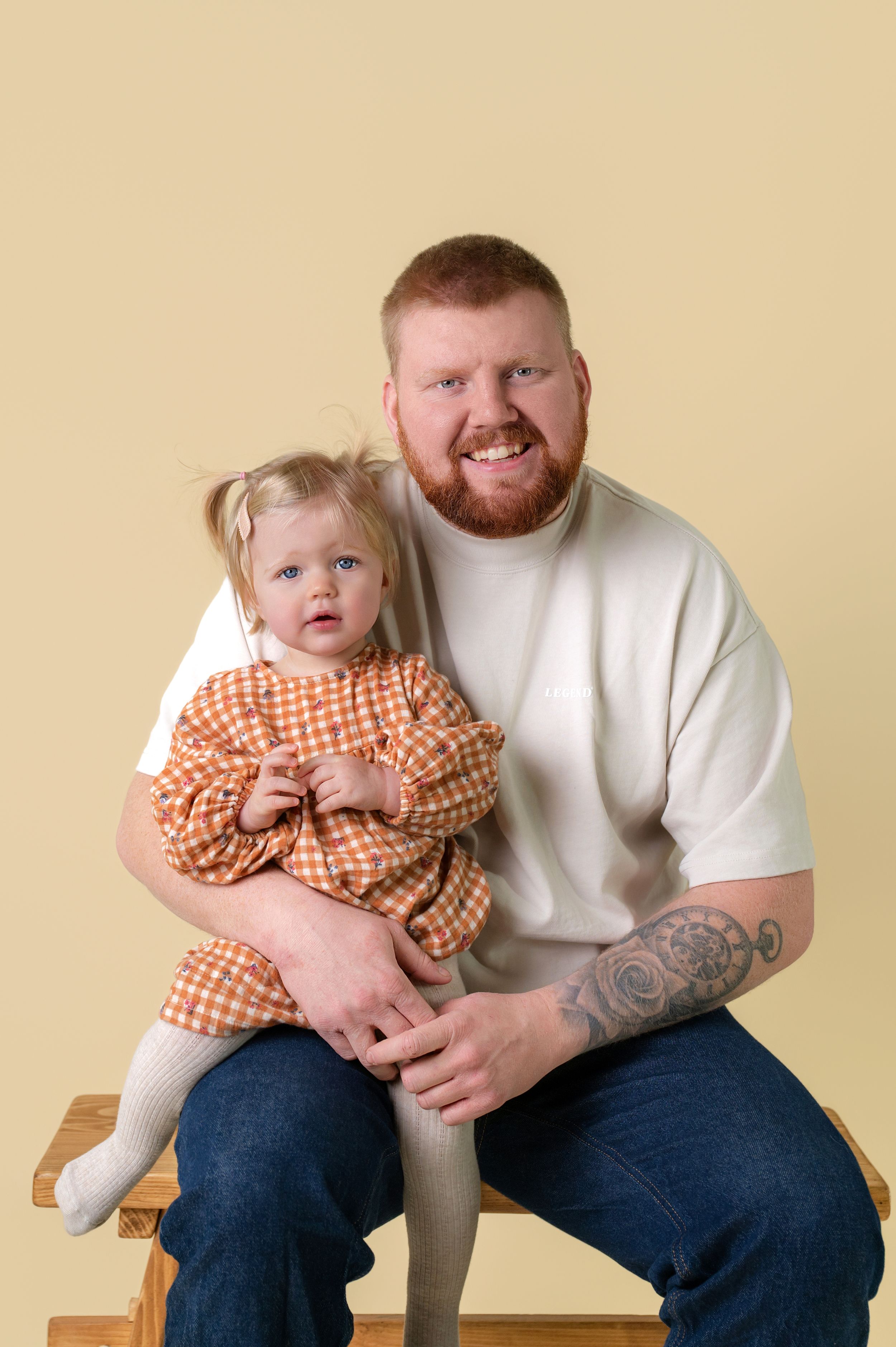 A parent and young child in orange gingham dress sit together for a portrait against a beige background.
