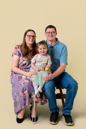 A family of three sits together on a bench wearing coordinated outfits in a studio setting.