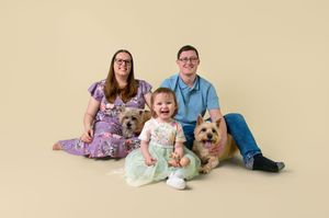 A family sits together on a beige backdrop with their small tan dog for a studio portrait.
