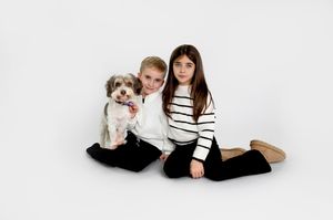 A small white dog sits with people on a black blanket in a minimalist studio setting.