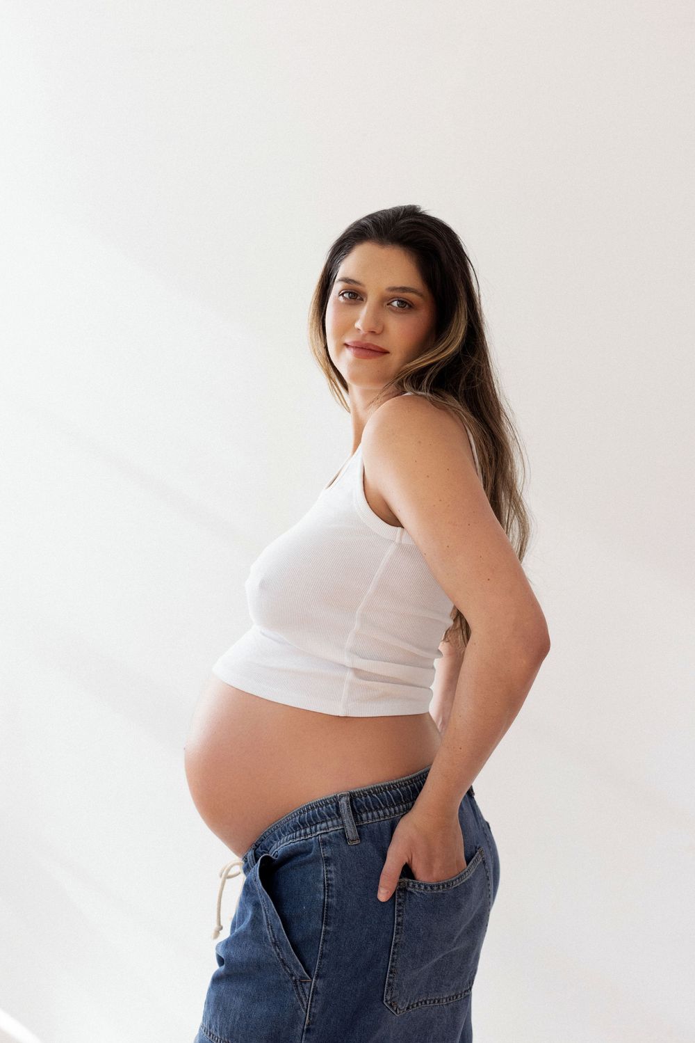 Pregnant woman in white crop top and jeans poses against white wall showing baby bump in maternity photo.
