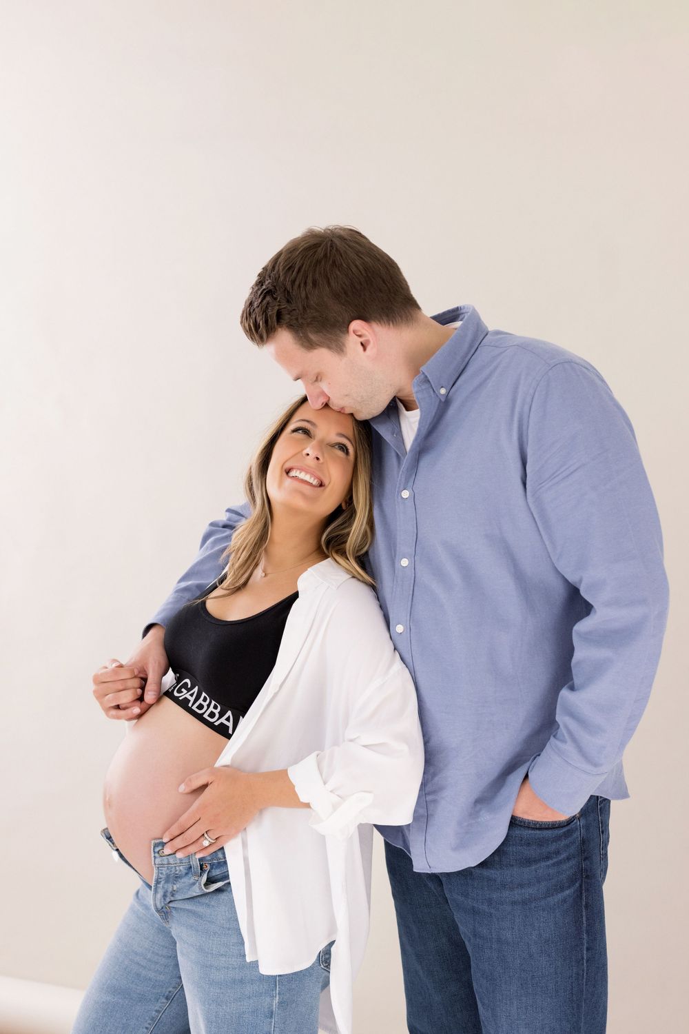 Expecting couple shares intimate moment in casual attire against white wall during maternity photoshoot.