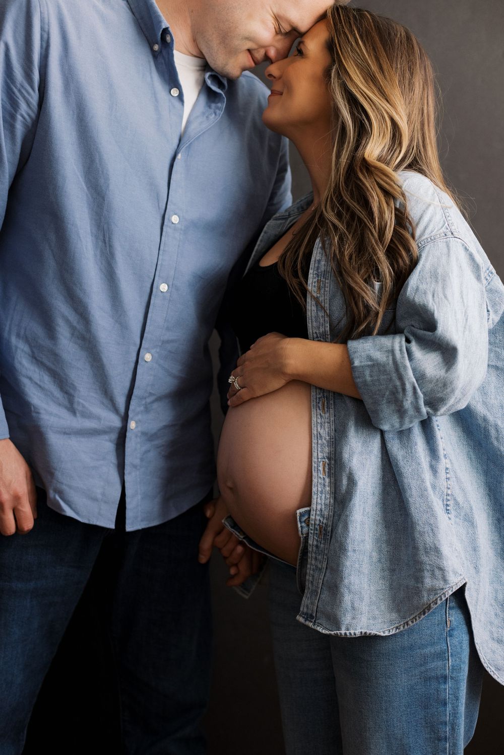 Intimate maternity photo of couple in matching denim and blue shirts against dark background.